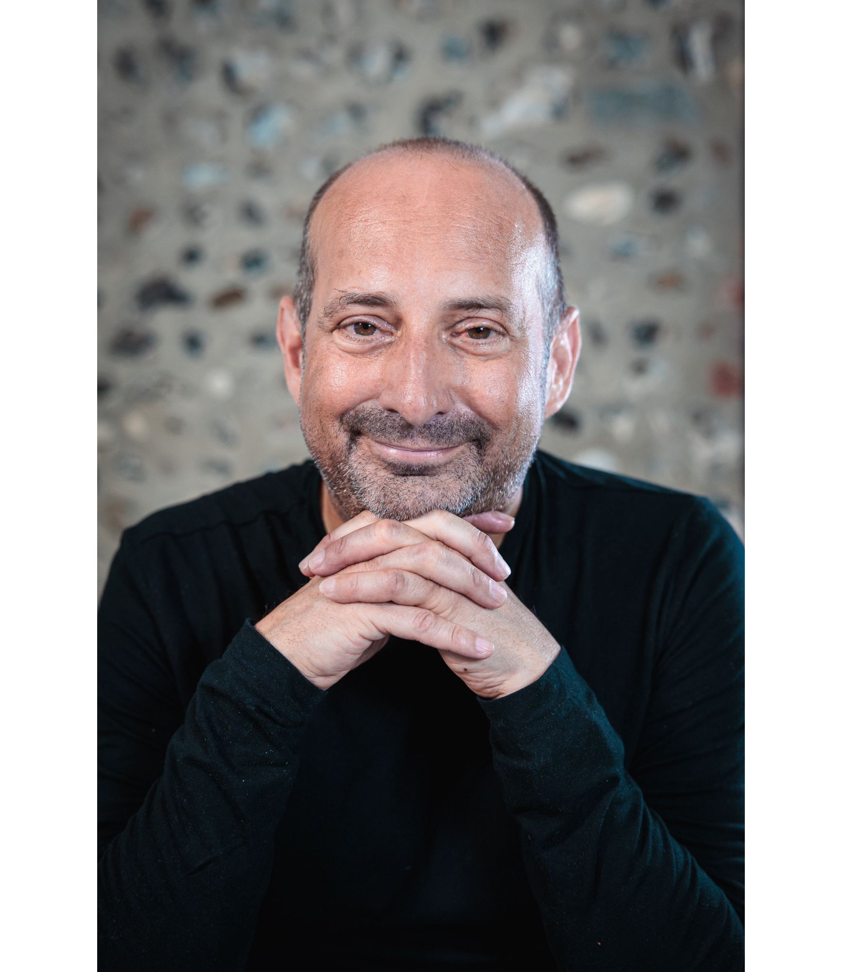 Clive has brown eyes, very little hair, a short beard and is wearing a black top and is smiling at the camera with his hands interlocked underneath his chin