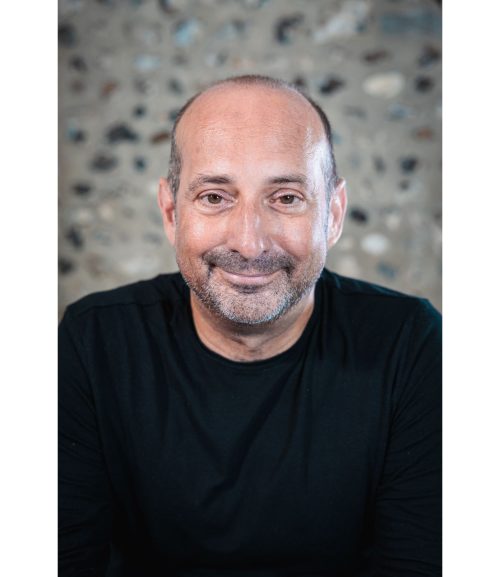 Clive has brown eyes, very little hair, a short beard and is wearing a black top and is smiling at the camera